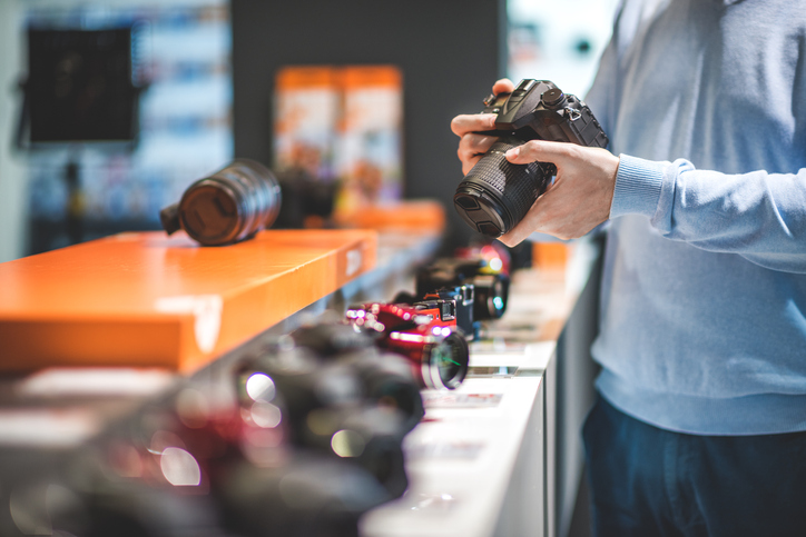 Unrecognizable male customer is holding black DSLR camera in the store. Row of other camera models is visible on the shelf.